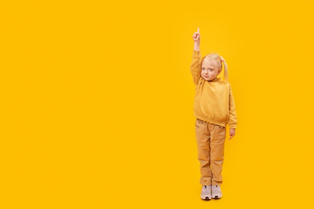 Preschool girl in yellow suit raised her hand and points finger upYellow backgroundCopy space for advertising mock up