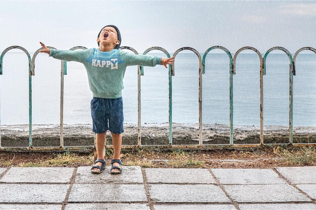 Preschooler boy spreading arms screams and enjoys on waterfront in rain