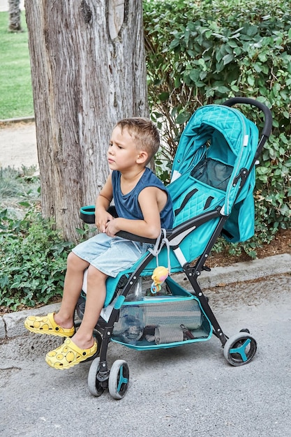 Preschooler frowns sitting in blue stroller of baby brother walking in park