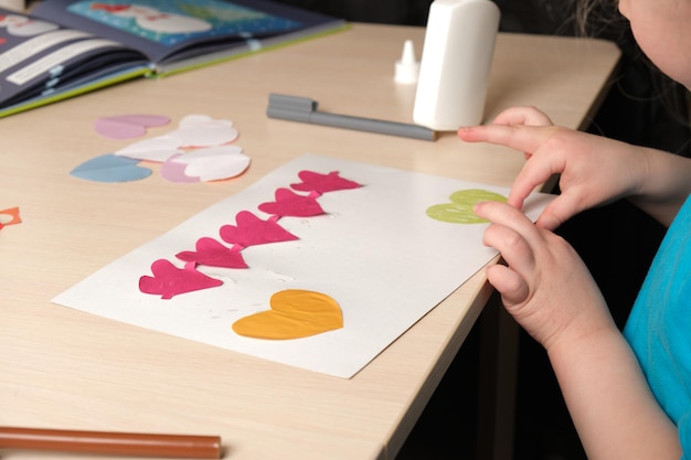 Photo preschooler girl in blue t-shirt makes applique sitting at the table, hearts cut out of colored bougue for card for valentine's day, children's creativity, atopic dermatitis on the hands of a child