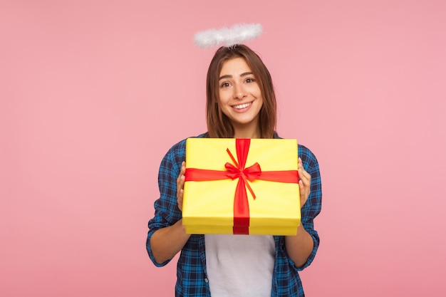 Present from angel Portrait of generous happy beautiful girl with halo over head holding gift box and smiling kindly congratulating on birthday indoor studio shot isolated on pink background