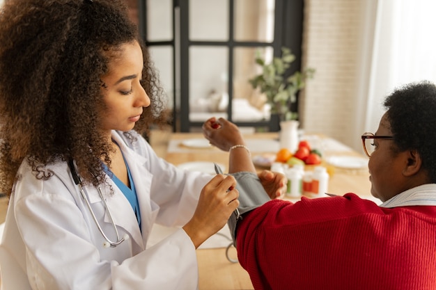 Pressure for woman. Curly dark-skinned nurse in white jacket measuring pressure for woman feeling bad