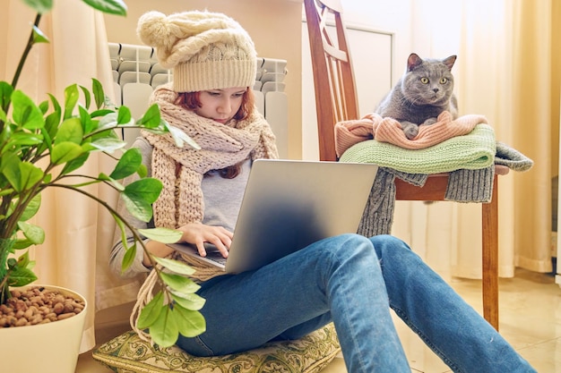 Preteen girl in scarf hat using laptop warming near heating radiator with pet gray cat