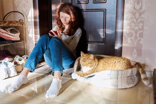 Preteen girl sitting on floor at home using smartphone with sleeping pet cat