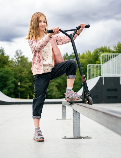 Preteen girl standing with scooter at street in the park and looking at camera cute child posing wit