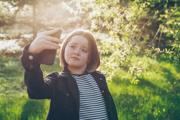 Preteen girl using camera of smartphone taking selfie recording video blog at park at spring day