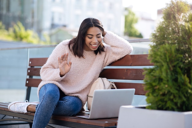 Pretty adorable pleasant woman sitting in park and talking to her mate from another country while using her laptop and free internet connection