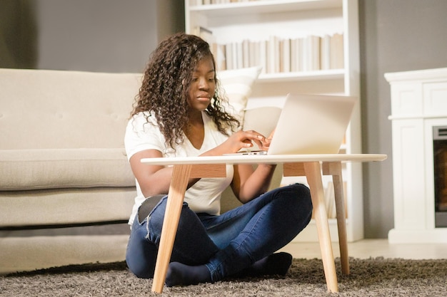 Pretty afro girl with folded legs sitting on the carpet and typing on her computer african girl look...