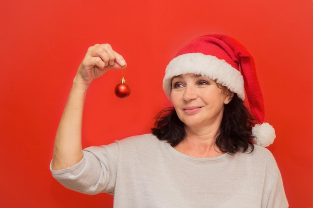 Pretty aged woman in santa hat holding christmas ball in her hands on red background