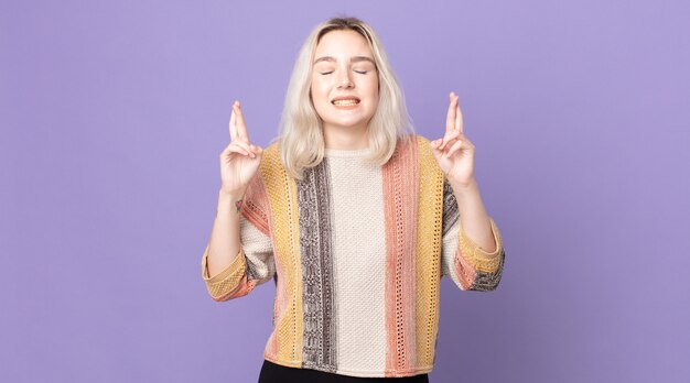 Pretty albino woman smiling and anxiously crossing both fingers, feeling worried and wishing or hoping for good luck