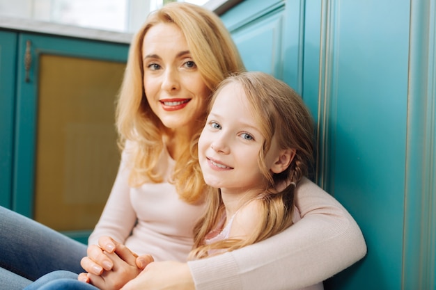 Pretty alert fair-haired mother and daughter smiling and sitting on the floor in the kitchen and cuddling