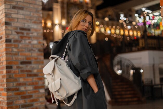 Photo pretty american young woman in a fashionable jacket with hood in stylish glasses with a stylish white backpack is walks on a shopping center. pretty girl fashion model enjoys the weekend. fash