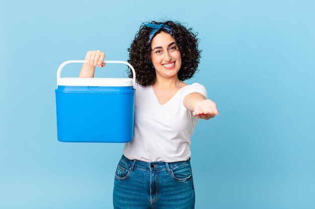 Pretty arab woman smiling happily with friendly and  offering and showing a concept and holding a portable refrigerator