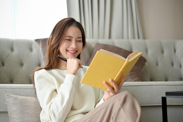 Pretty Asian female relaxes on a comfy carpet in the living room and reads her favourite novel