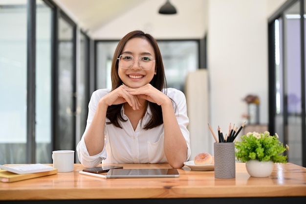 Pretty Asian female sits at her desk hands on chin smiling and looking at the camera