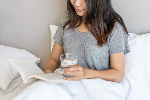 Pretty Asian woman sitting in bed reading a book while holding a glass of mineral water in the morning at home Relax and lifestyle in free day concept Closeup