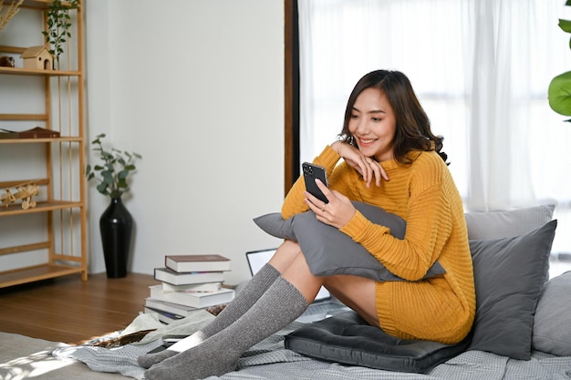 Pretty Asian woman in sweater and comfy socks using her mobile phone in her living room