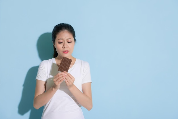 pretty attractive girl standing on blue wall background and looking at chocolate cookie thinking tooth decay problem hesitating whether to eating.