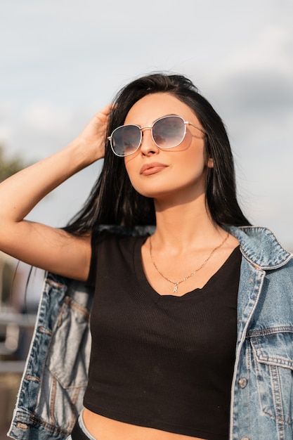 Pretty attractive young hipster woman in black top in a denim blue jacket in stylish black sunglasses relaxes near a vintage wooden wall. Sexy brunette girl model in trendy clothes rests outdoors.