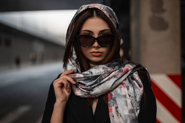 Pretty attractive young woman fashion model in elegant stylish shawl in sunglasses in black coat posing in the city on the street. Fashionable business girl outdoors on spring day. Beauty modern lady.