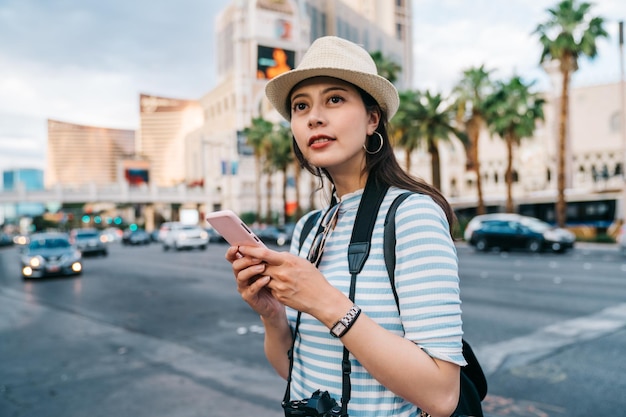 pretty backpacker standing on the busy road and searching the right direction by cellphone. young girl using communicate app by smartphone. elegant woman enjoy the beauty of the blue sky.