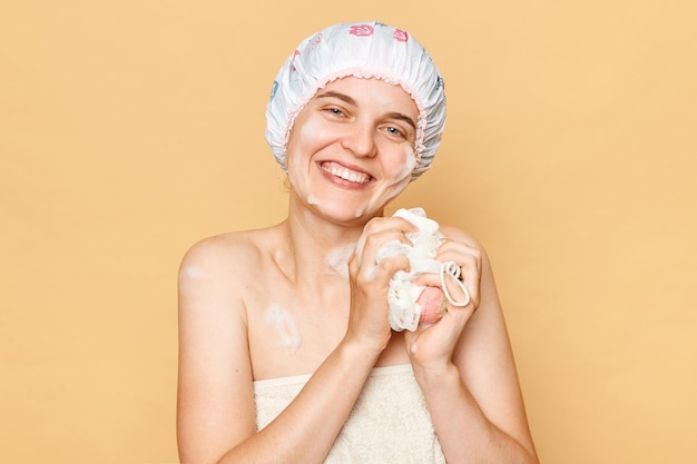 Pretty beautiful woman wearing shower cap standing isolated over beige background looking at camera holding sponge enjoying freshness after taking bath in the morning