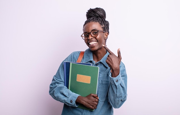 Pretty black woman smiling confidently pointing to own broad smile. student with books concept