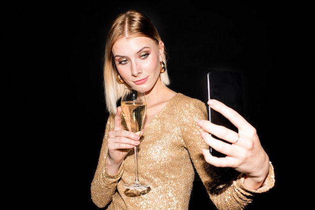 Pretty blond girl with flute of champagne and smartphone making selfie while holding drink by her chest and looking at camera