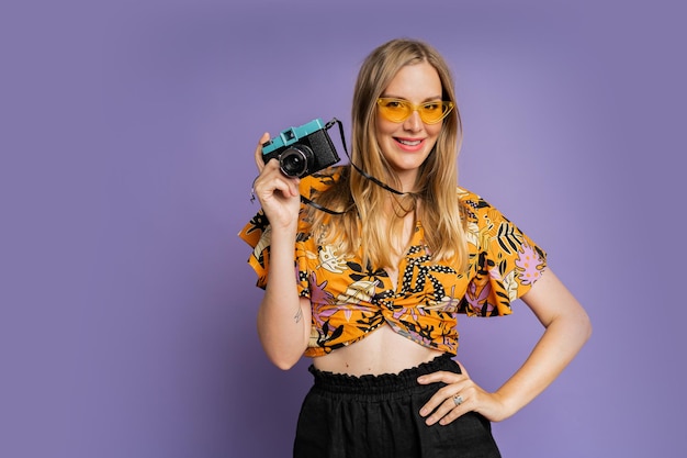 Pretty blond woman in stylish summer outfit holding camera and posing on purple background in studio Travel and summer fashion concept