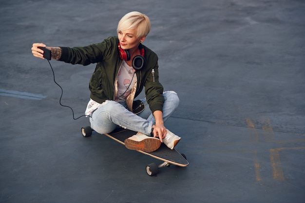 Pretty blond woman with headphones, making self photo on phone while sitting on the longboard at sunset.