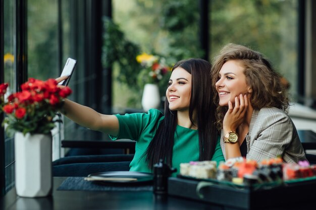 Photo pretty blonde and brunette girl taking a picture by the cell phone with sushi plate on table. chenese eat, friends.