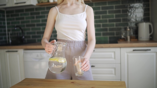 Pretty blonde drinks lemon water. Young woman pours water with lemon into a glass and drinks in the kitchen. Slow motion, camera moves from bottom to top, HD.