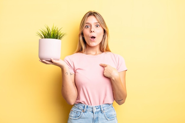 Pretty blonde girl looking shocked and surprised with mouth wide open, pointing to self. houseplant concept