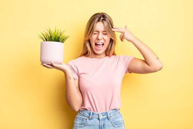 Pretty blonde girl looking unhappy and stressed, suicide gesture making gun sign. houseplant concept