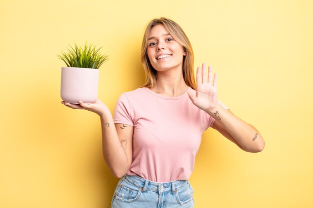 pretty blonde girl smiling happily, waving hand, welcoming and greeting you. houseplant concept