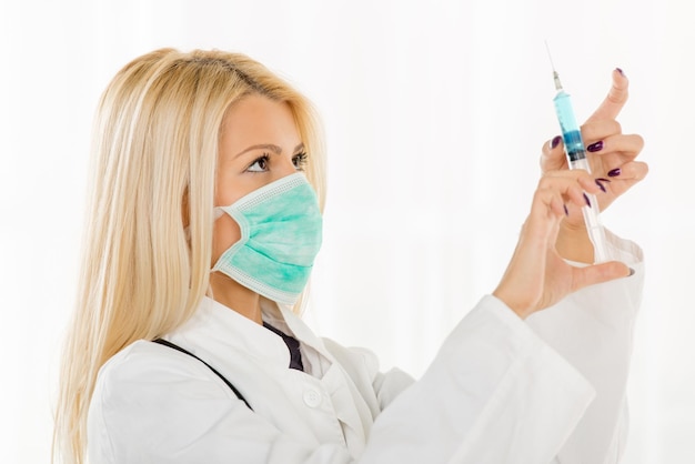 Pretty blonde young woman in a white coat, with a stethoscope over the neck holding and preparing an injection.