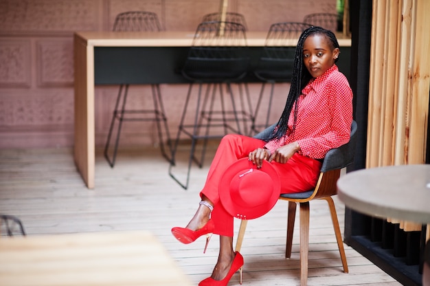 Pretty braids business african american lady bright bossy person friendly wear office red shirt, hat and trousers, sit on chair.