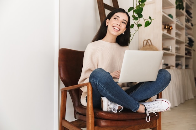 pretty brunette woman resting in apartment and working on laptop computer while sitting on chair