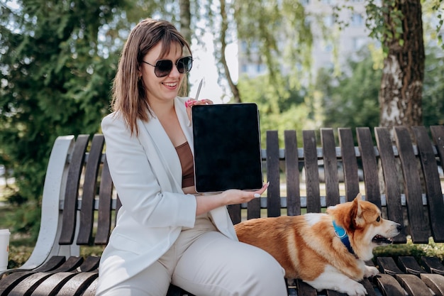 Pretty businesswoman in white suit sitting in city parkland with Welsh Corgi Pembroke dog and demonstrate digital tablet xA