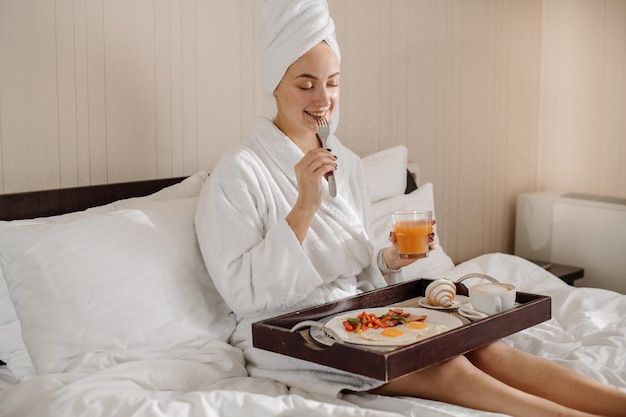 Pretty caucasian female in bathrobe and towel on head having a breakfast in bed in the morning at the hotel room