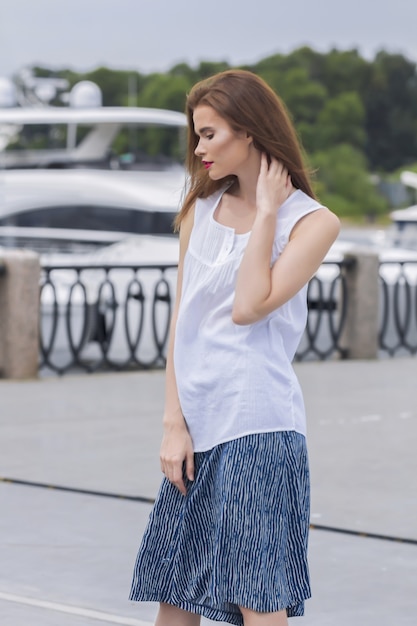 Pretty caucasian girl spending time at the pier in front of luxury yachts in a light blouse and blue divided skirt.