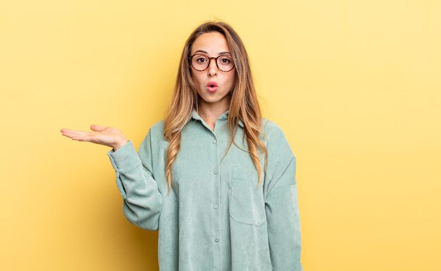 Pretty caucasian woman looking surprised and shocked, with jaw dropped holding an object with an open hand on the side