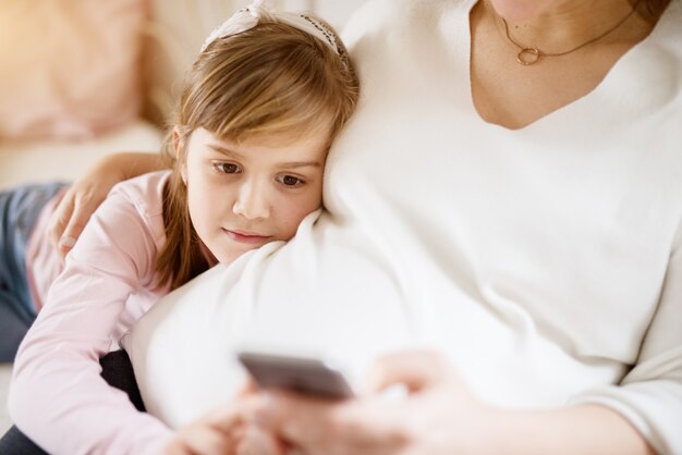 Pretty cheerful little toddler girl lying on the belly of her pregnant mother while using mobile. Family and new life concept.