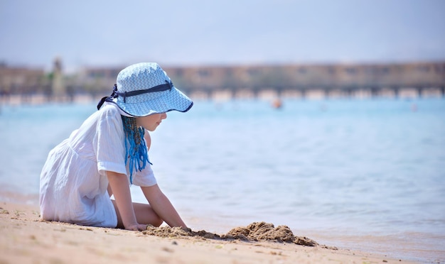 Pretty child girl in big hat and white dress playing with wet sand on background of blue sky and clear ocean lagoon water