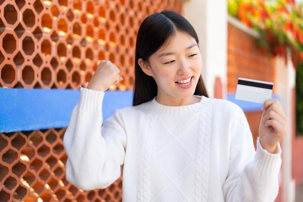 Pretty Chinese woman holding a credit card at outdoors celebrating a victory