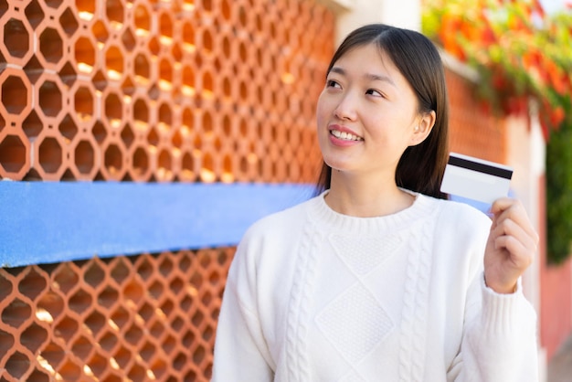 Pretty Chinese woman holding a credit card at outdoors looking up while smiling