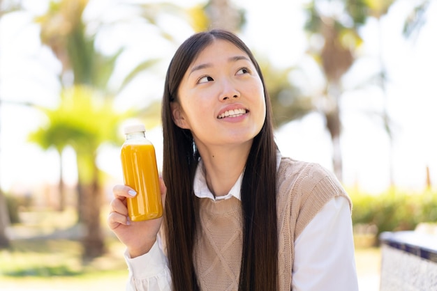 Pretty Chinese woman holding an orange juice at outdoors looking up while smiling