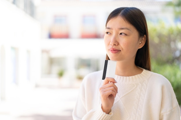 Pretty Chinese woman at outdoors holding a credit card and thinking