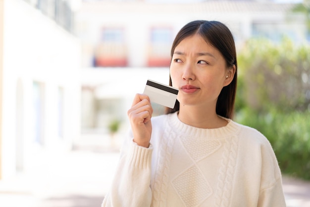 Pretty Chinese woman at outdoors holding a credit card and thinking