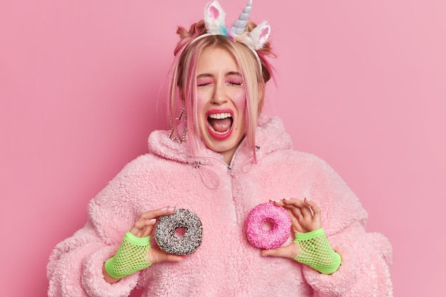 Photo pretty emotional woman exclaims loudly feels extremely happy holds two glazed donuts keeps mouth wide opened wears unicorn headband and fur coat